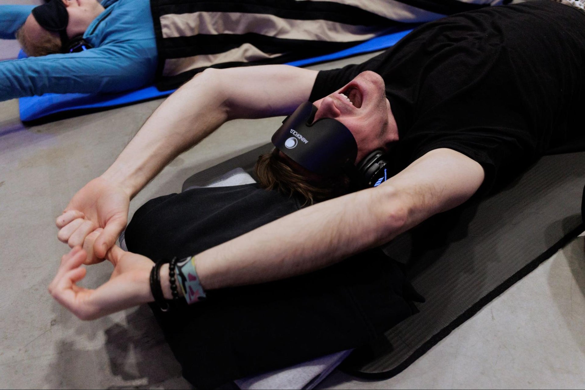 A man wearing a blindfold and headphones stretches his arms deeply while lying down during a breathwork session.