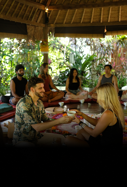 Participants seated cross legged in an open pavilion surrounded by greenery, exchanging cups during a group ceremony