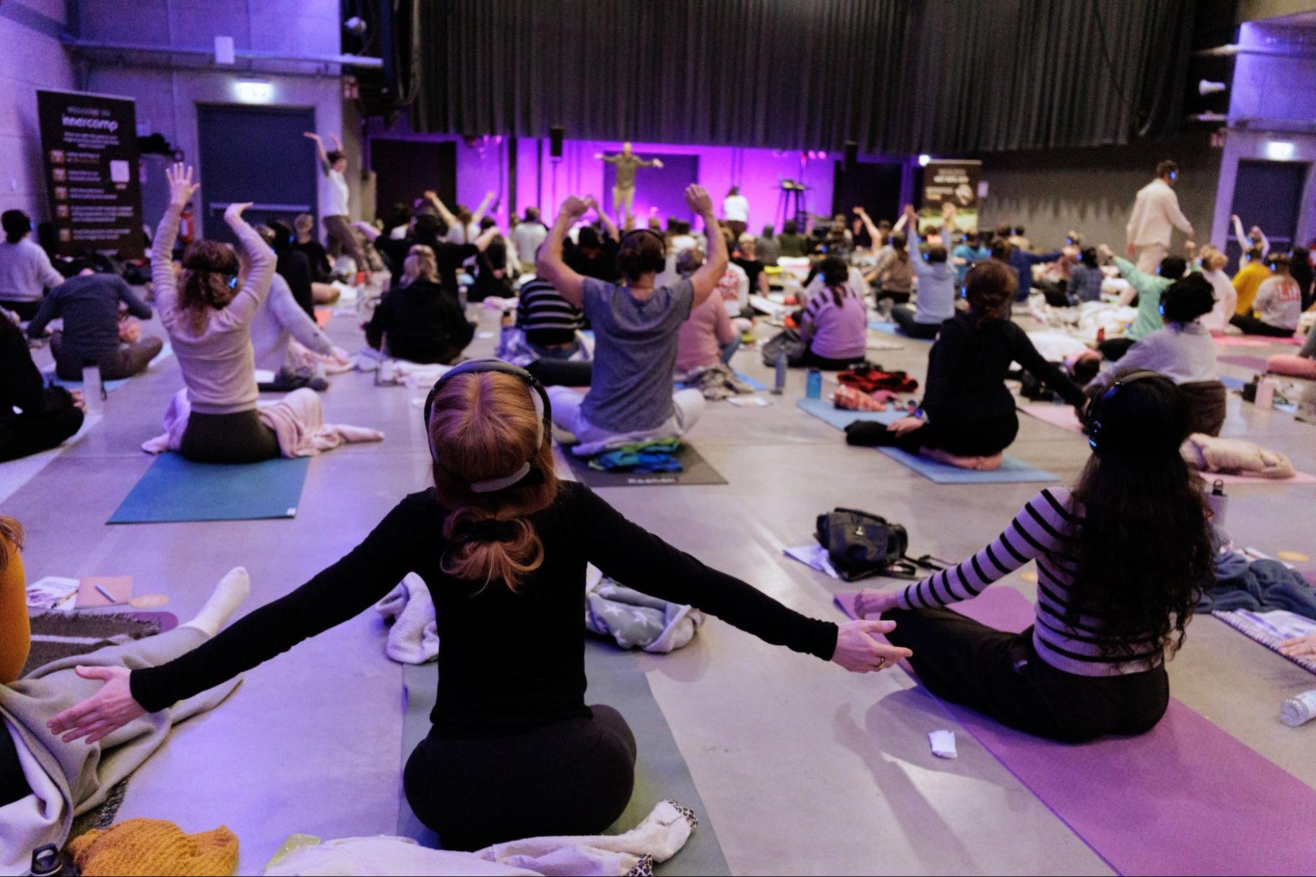 Large group of participants sitting on mats with arms raised during a guided breathwork workshop under purple lighting