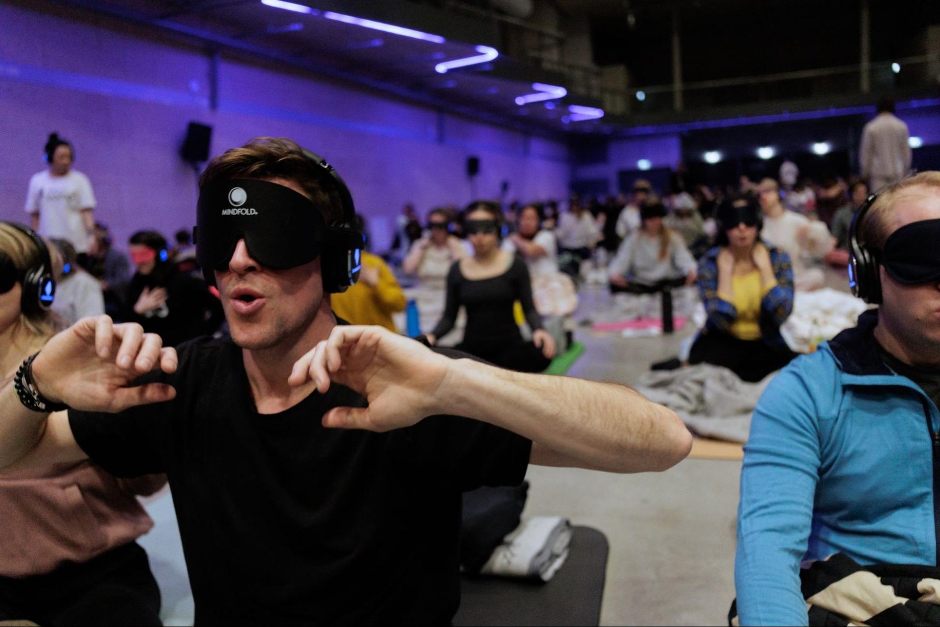 A man in a blindfold and headphones raises his hands expressively during a guided group breathwork session.