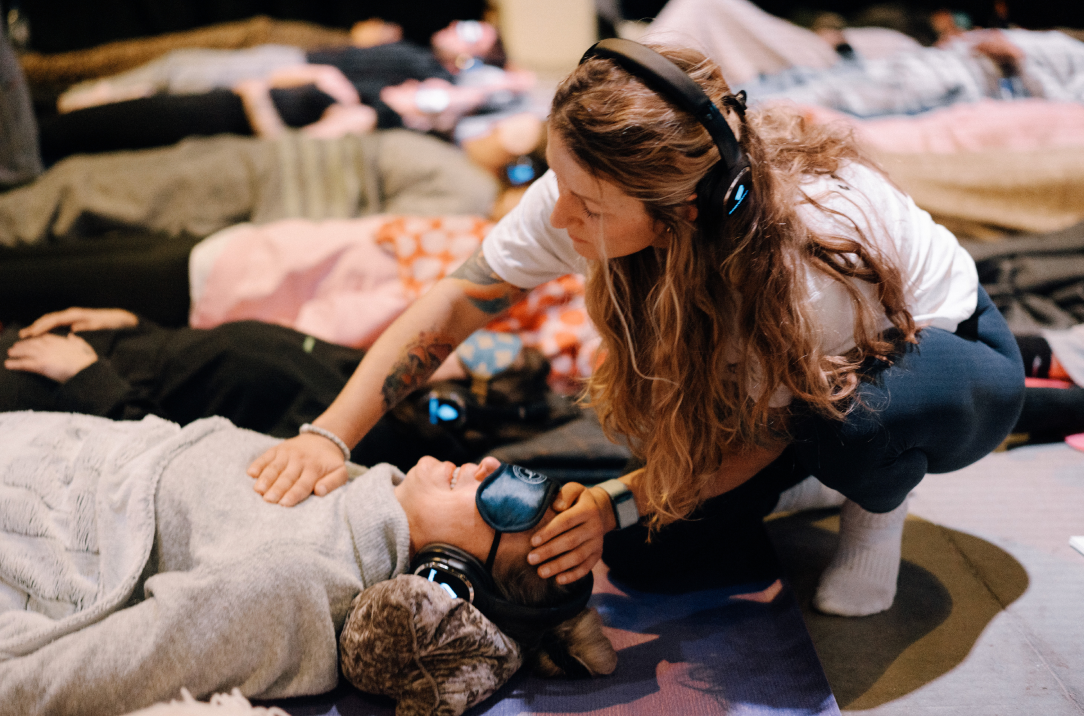Woman acting as a sitter, placing a hand on a breather's chest while they lie down wearing headphones during breathwork